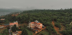 Um edifício com vários andares e telhado vermelho, cercado por vegetação densa e um caminho de terra.