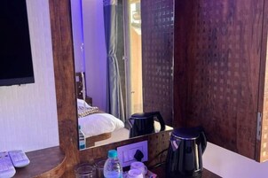 Habitación de hotel con un escritorio de madera, un espejo, una cama, una silla negra, una tetera y una botella de agua.