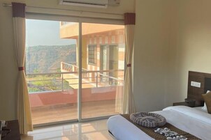 Habitación de hotel con cama, una cama para mascotas, mesita de noche, ventana con cortinas y vistas a un edificio y paisaje.