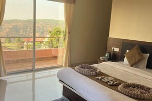 Quarto de hotel com uma janela grande, uma cama com duas caminhas para animais de estimação, uma mesa de cabeceira com abajur e vista para uma paisagem montanhosa.