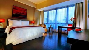 A hotel room with a bed, a desk, a chair, a small table, and a large window with curtains. - 100 Sunset Kuta Hotel & Ballroom (Bali)