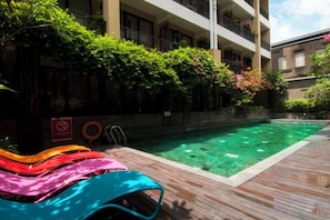 A swimming pool with a wooden deck, lounge chairs, and a building with balconies in the background. - 100 Sunset Kuta Hotel & Ballroom (Bali)