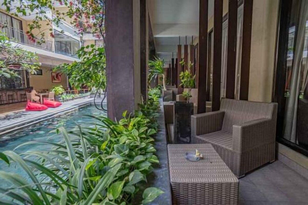 Zona junto a la piscina con un sofá y una mesa de mimbre, rodeada de exuberante vegetación y una piscina cristalina.