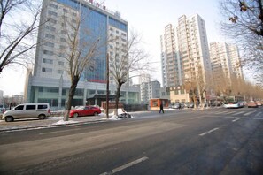 En gata med höga byggnader, parkerade bilar och kala träd.