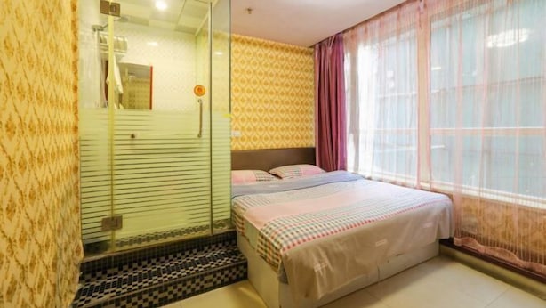 A hotel room with a glass shower enclosure, a bed with a patterned bedspread, and a large window with sheer curtains.