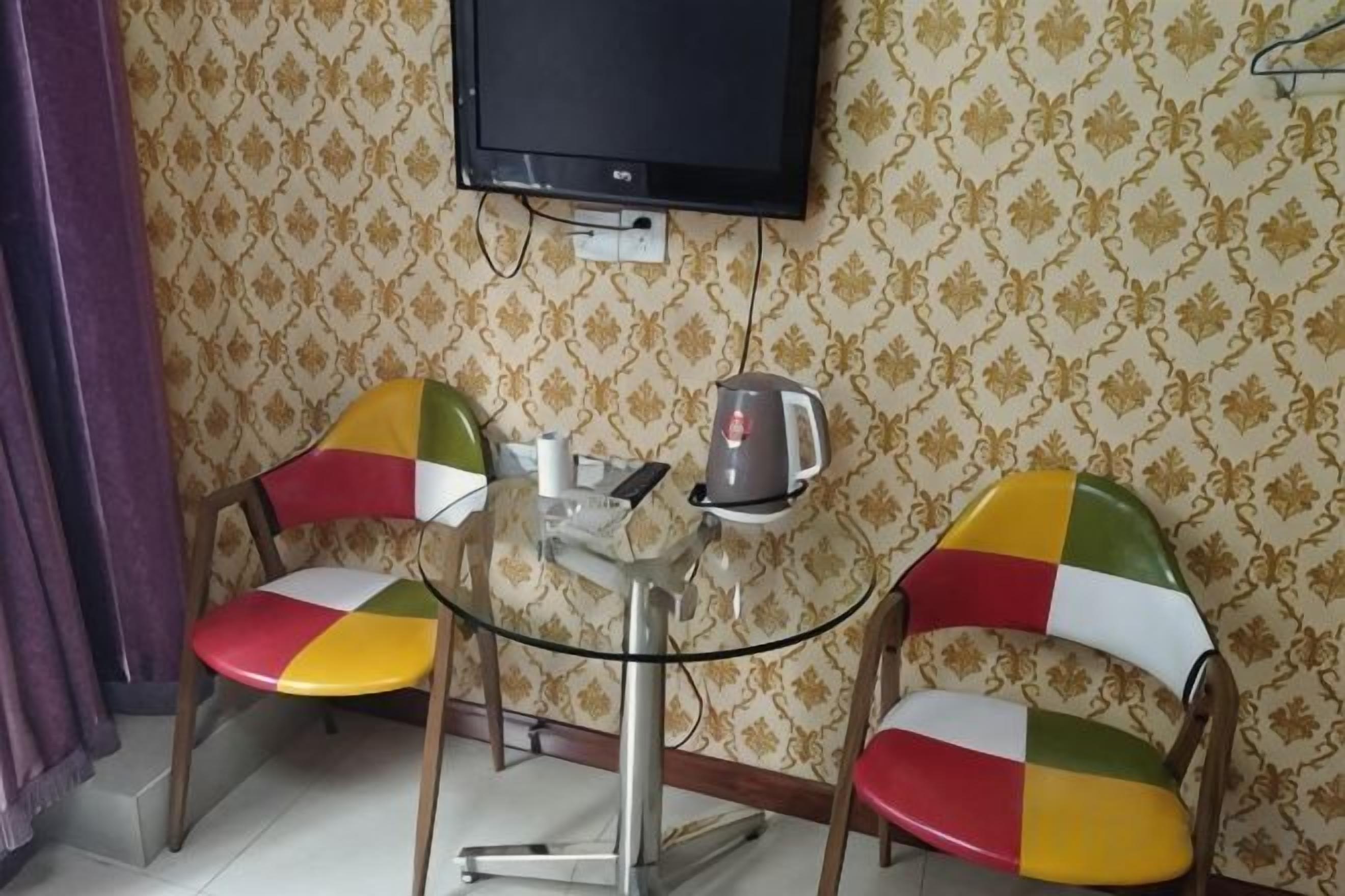Una habitación pequeña con dos sillas coloridas, una mesa de vidrio, una tetera y un televisor montado en la pared.