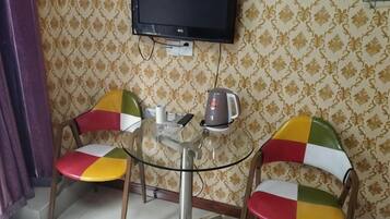 Una habitación pequeña con dos sillas coloridas, una mesa de vidrio, una tetera y un televisor montado en la pared.