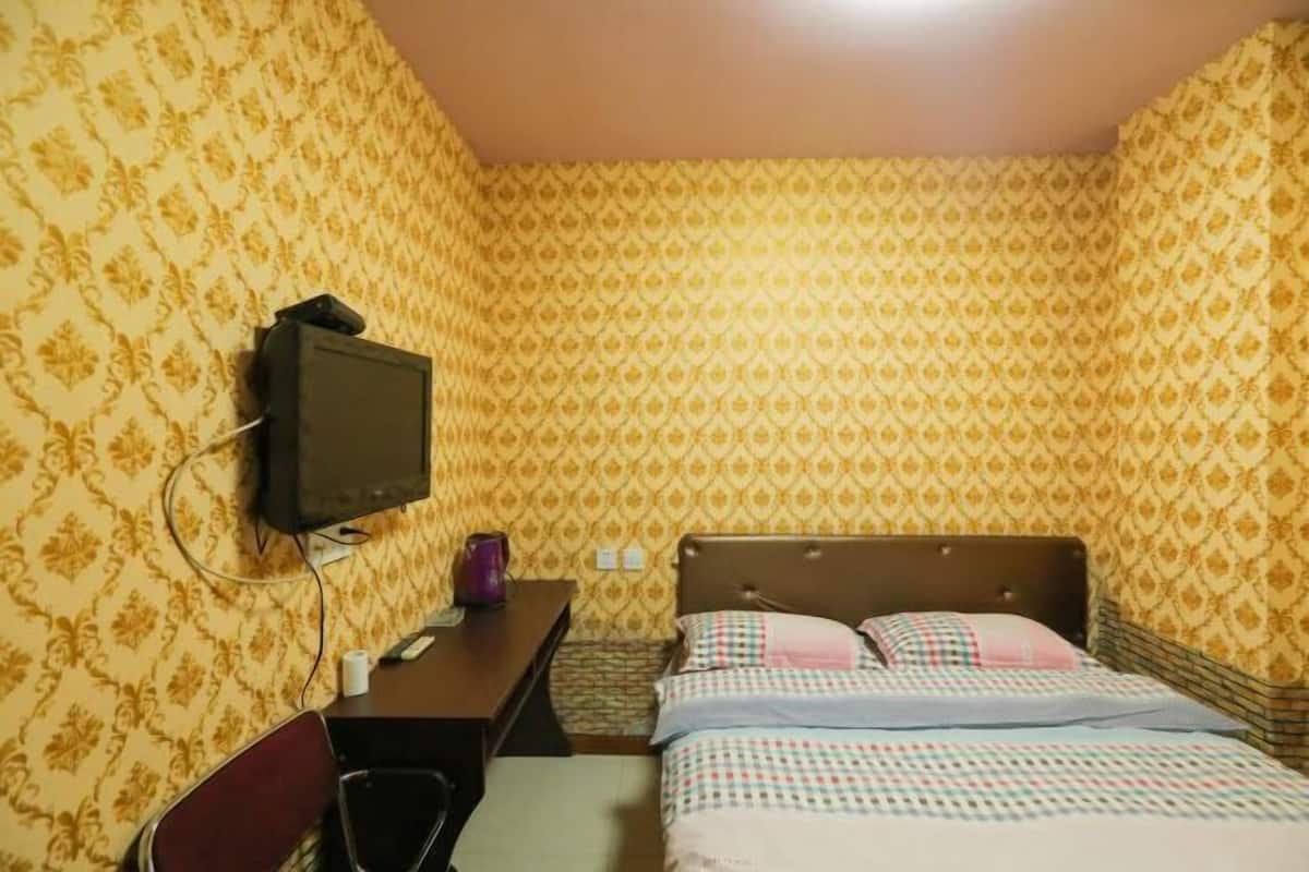 A small, single-room hotel room with a bed, a desk, a chair, and a television mounted on the wall.