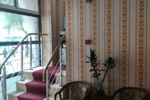 Una escalera con barandilla que sube a un piso superior, pared con diseño y dos sillas con una planta en maceta.