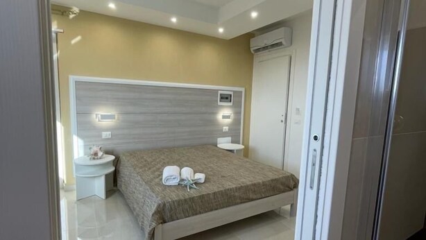 A modern bedroom with a large bed, bedside tables, an air conditioning unit, and a small round table with a lamp. - B&B Jerocades (Parghelia)