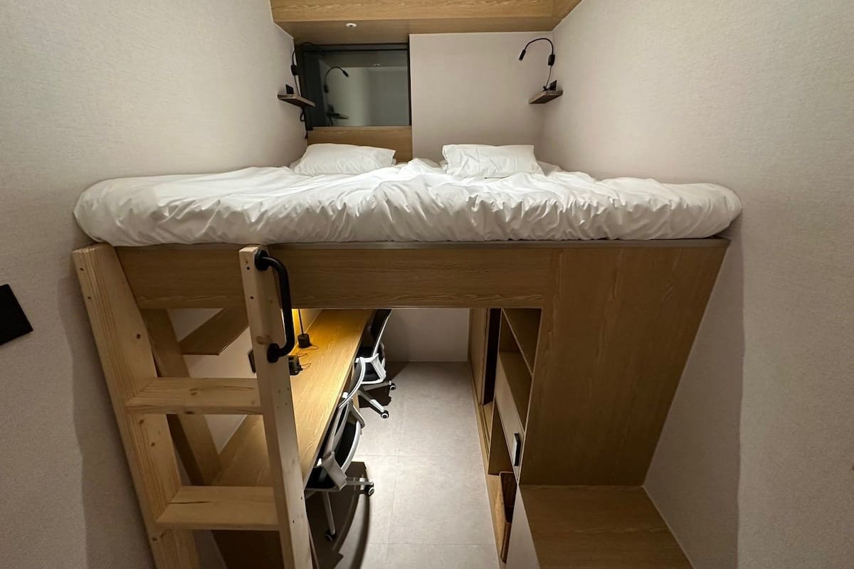 A compact room with a bunk bed, a small desk with a chair, and a shelf with a lamp.
