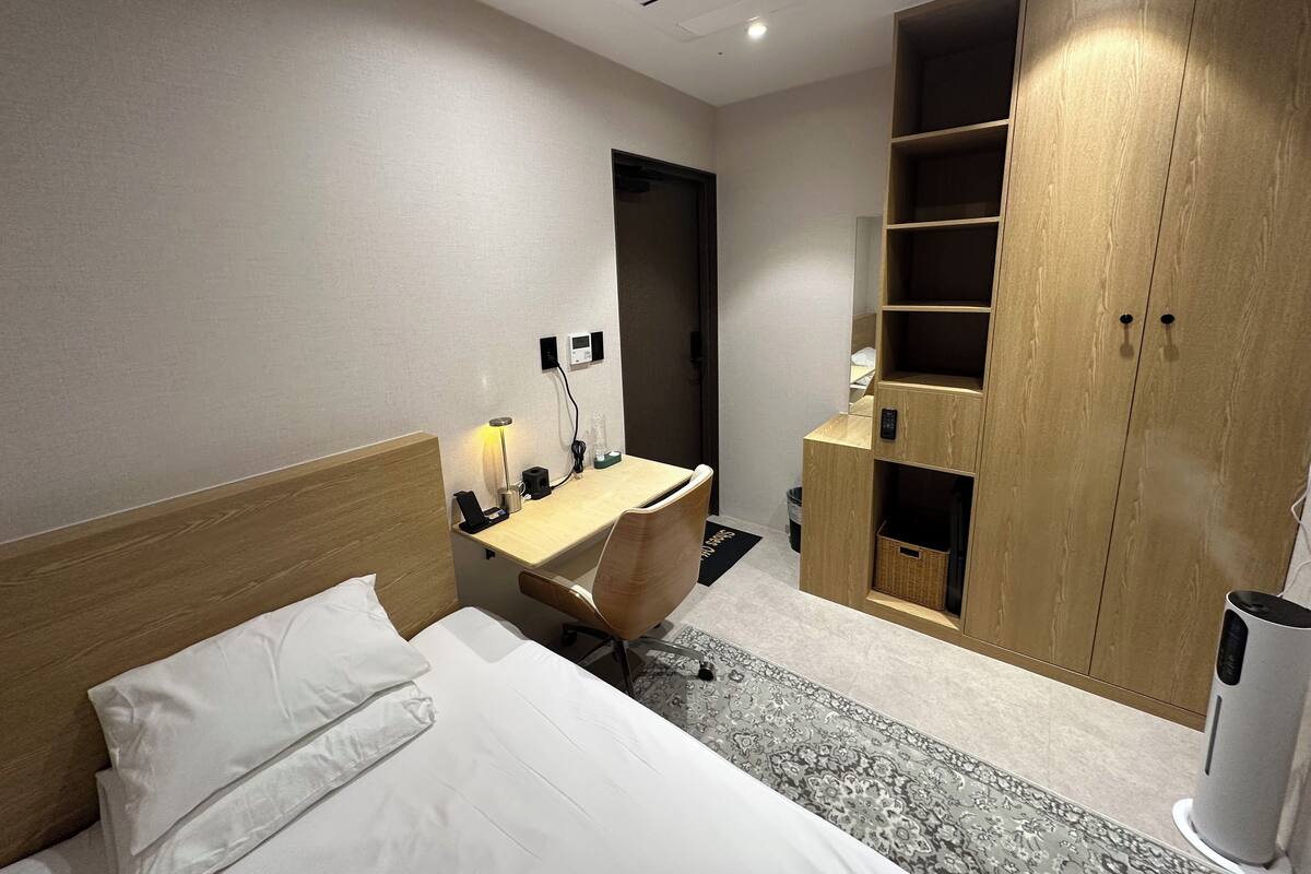 A hotel room with a bed, a desk, a chair, a wardrobe, and a trash bin.