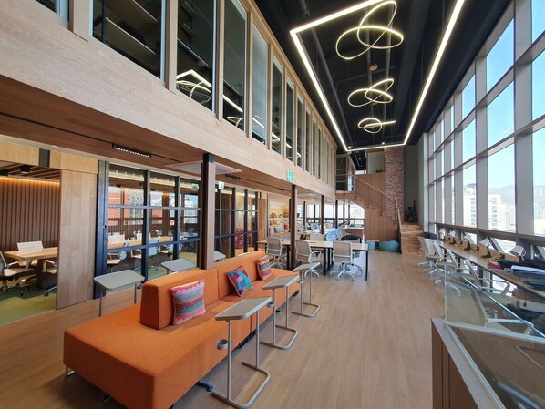 A modern library with wooden floors, orange seating, and large windows. - NOMADX Stay Hostel (Busan)
