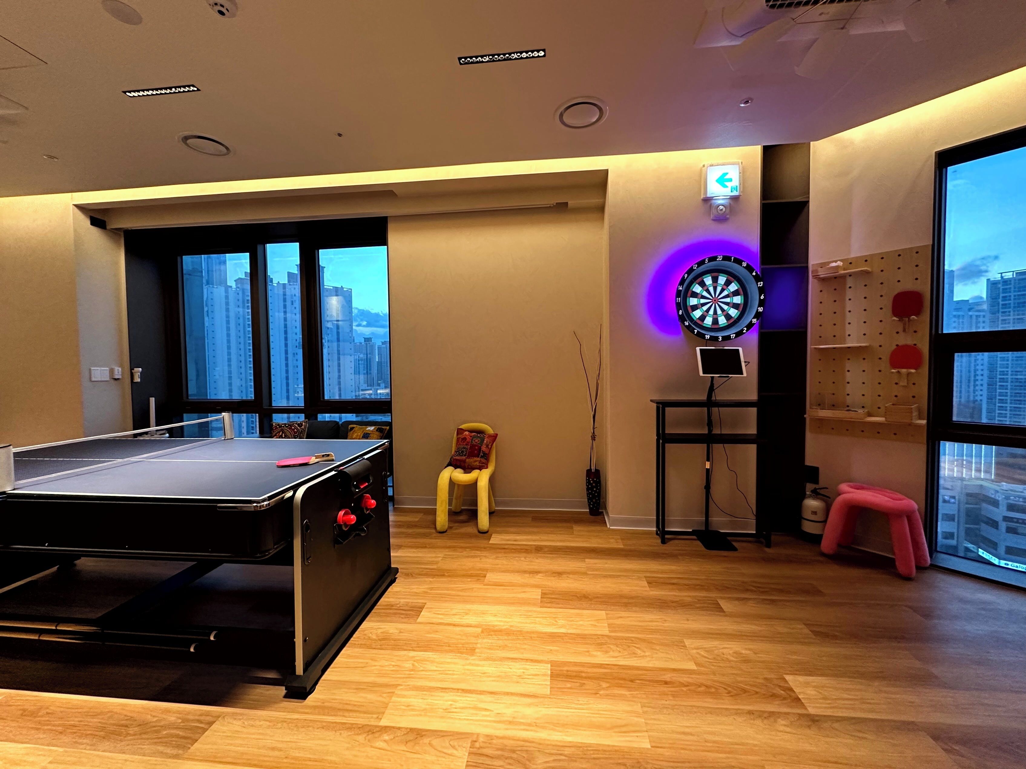 A room with a pool table, a dartboard, a small table with a monitor, and a window with a view of tall buildings.