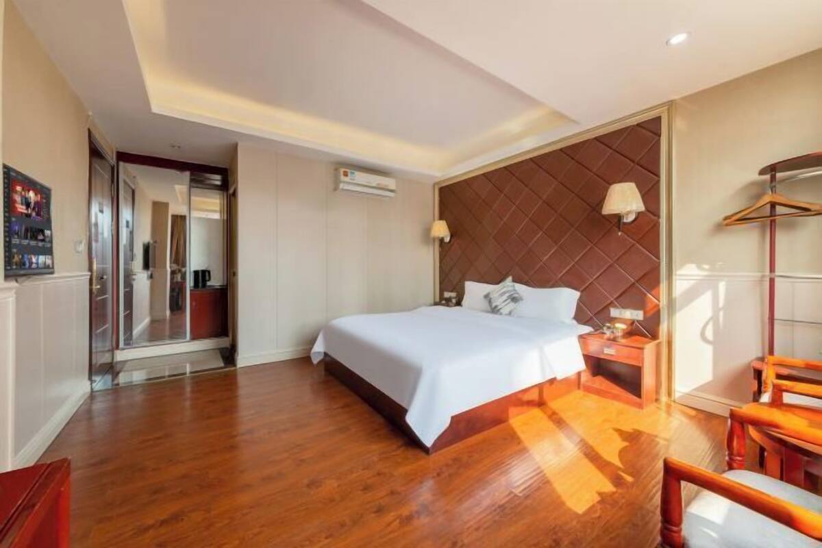 A hotel room with a bed, bedside table, lamp, wooden floor, and a mirror.