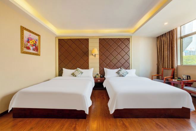 A hotel room with two beds, a wooden floor, a wall-mounted lamp, and a window with curtains.