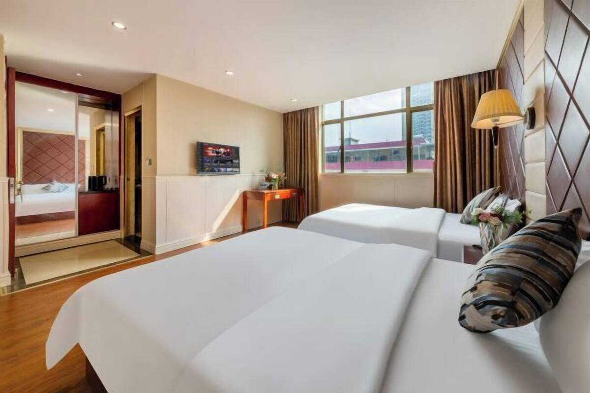 A hotel room with two beds, a desk, a TV, and a window with curtains.