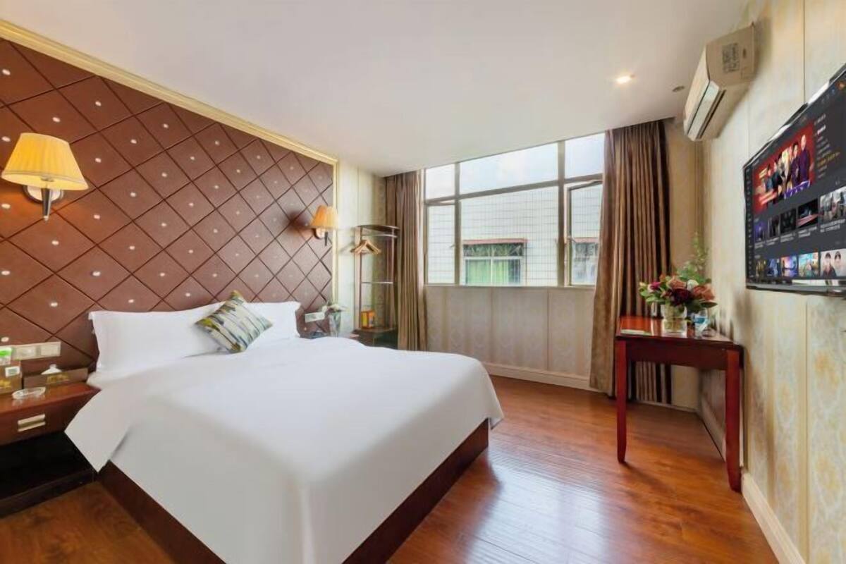 A hotel room with a large bed, a TV on the wall, a window with curtains, and a wooden floor.