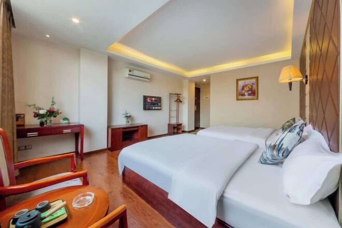 A hotel room with a large bed, a desk with a chair, a small table, and a wall-mounted lamp.
