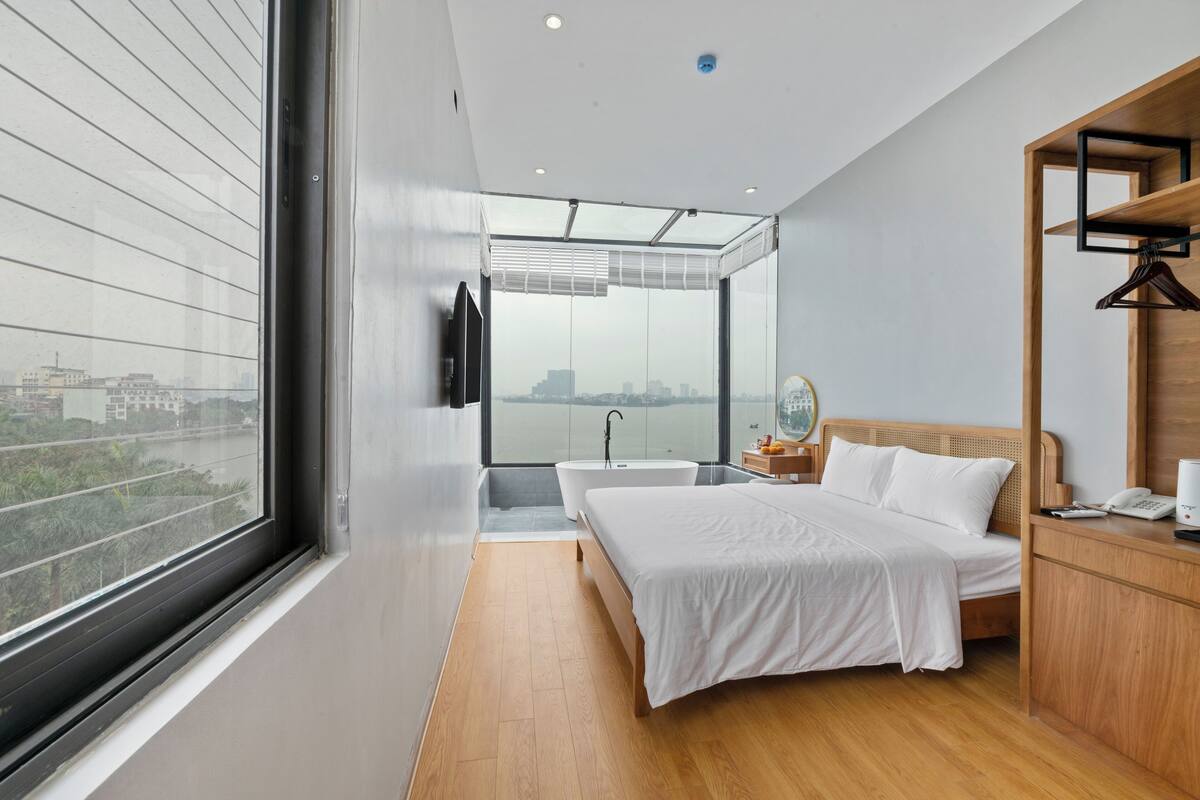 A modern bedroom with a large window, a bathtub, a bed, and a wooden nightstand.