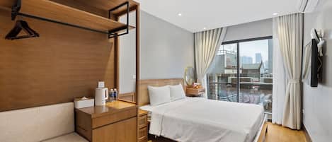 A hotel room with a wooden bench, a bed with white bedding, a bedside table with a lamp, and a view of the city through large windows.