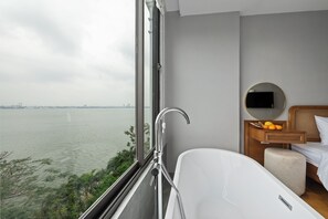 Une salle de bain moderne avec une baignoire, une table de chevet en bois ornée d’oranges, et une vue sur une étendue d’eau.