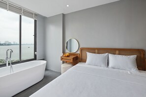 A modern hotel room with a large bathtub, a wooden bedside table, a round mirror, and a bed with white linens. - Lakeview Residence (Hanoi)