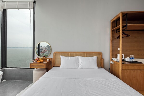 A hotel room with a large bed, a wooden headboard, a bedside table with fruit, a round mirror, and a view of the water. - Lakeview Residence (Hanoi)