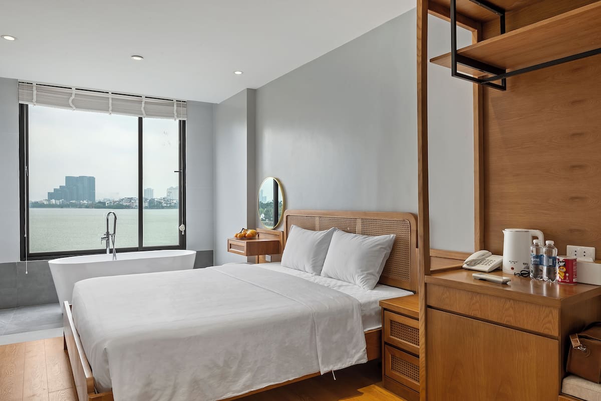 A modern hotel room with a large bed, a wooden nightstand, a mirror, and a view of a cityscape through large windows.