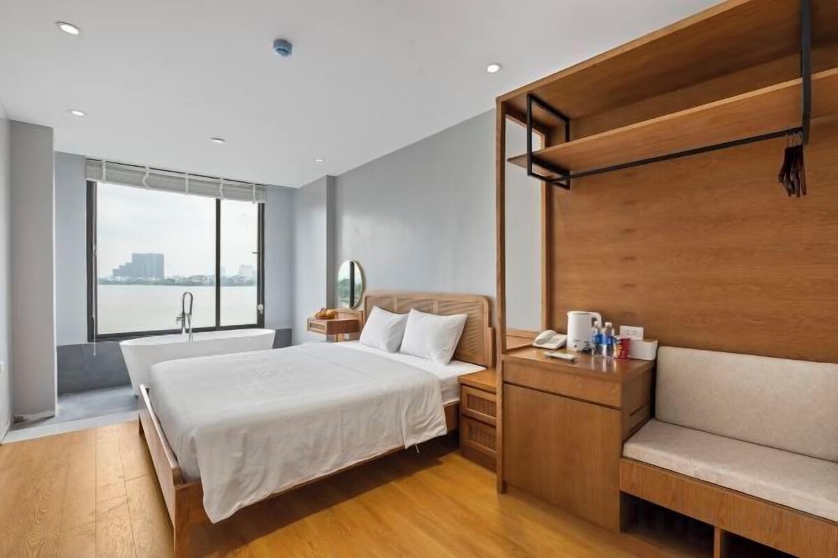 A hotel room with a large bed, a wooden bench, a desk with a chair, and a view of a body of water.