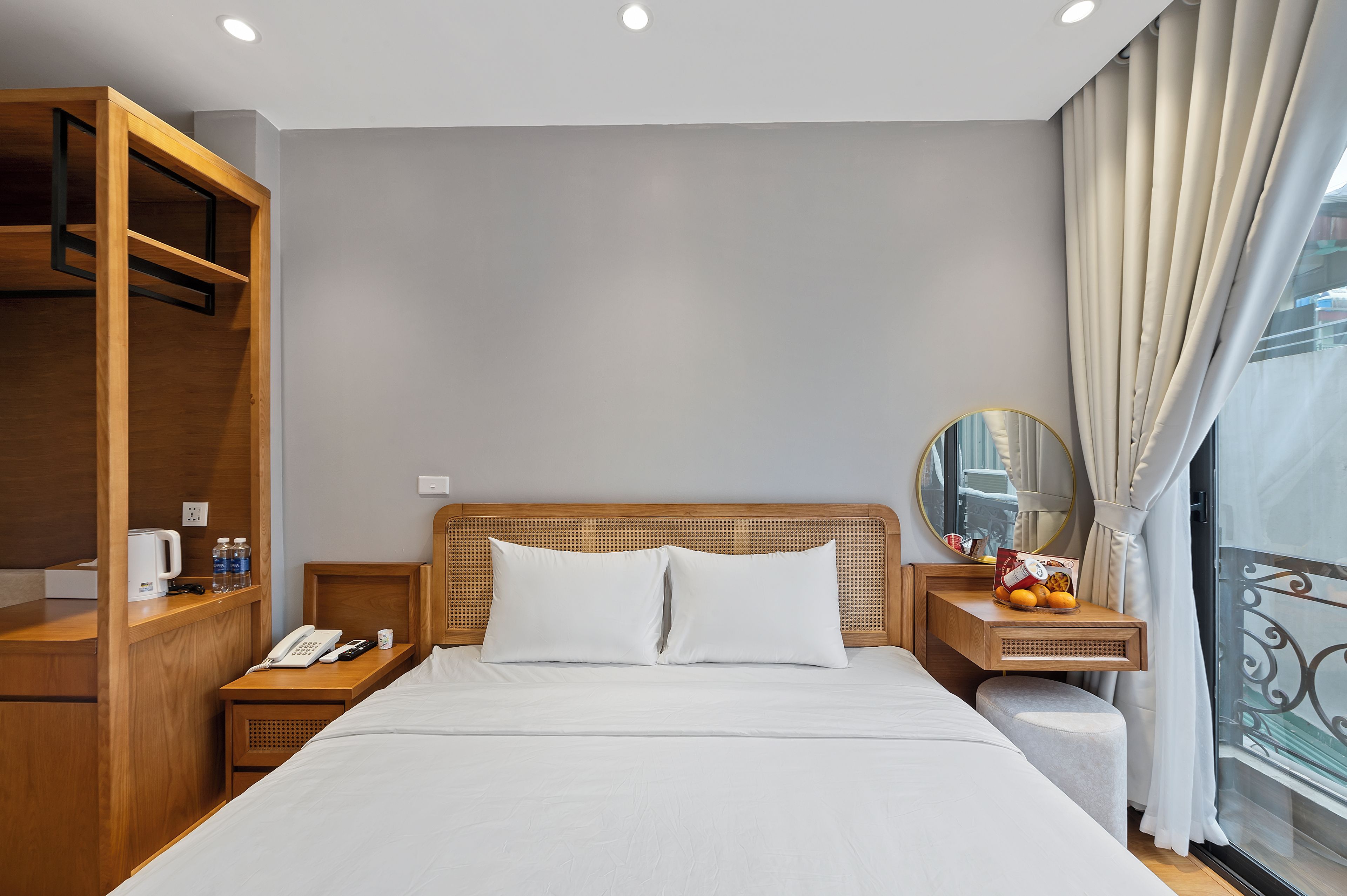 Habitación de hotel con cama, cabecero de madera, mesita de noche con teléfono, una pequeña mesa con espejo y una ventana con cortinas.