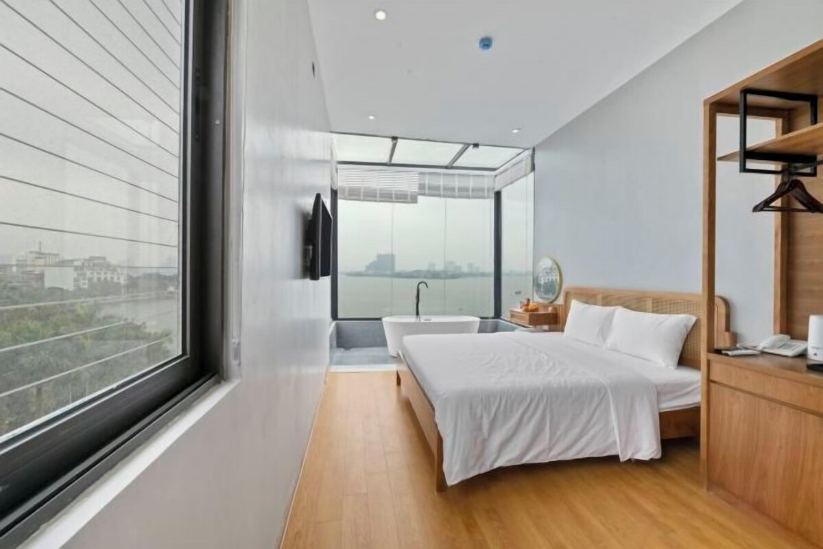 A modern hotel room with a large window, a bed, a bedside table, and a wooden wardrobe.