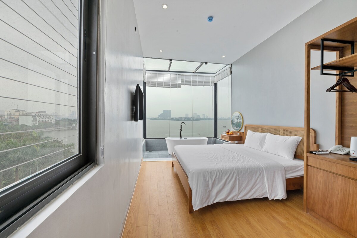 A modern bedroom with a large window, a bathtub, a bed, and a wooden nightstand.