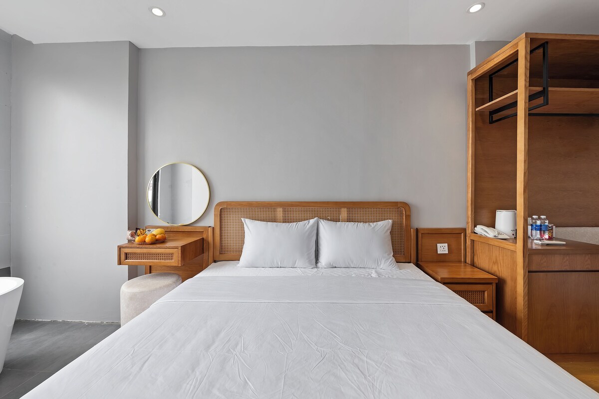 A modern hotel room with a large bed, a wooden headboard, a bedside table with a lamp, a small wooden cabinet, and a round mirror on the wall.