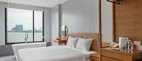 A modern hotel room with a large bed, a wooden nightstand, a mirror, and a view of a cityscape through large windows.