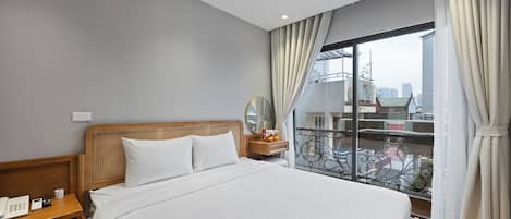 A hotel room with a large bed, wooden headboard, bedside table with a phone and remote controls, a nightstand, and a view of the city through a balcony with metal railings.