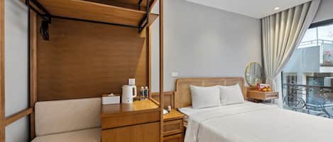 Habitación de hotel con un banco de madera, una cama con sábanas blancas, una mesita de noche con una tetera y vistas a un balcón con barandilla.