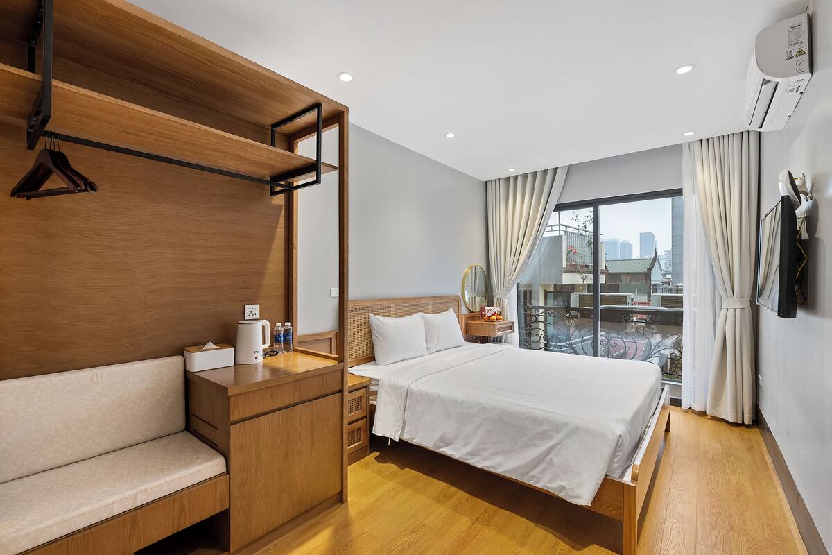 A hotel room with a wooden bench, a bed with white bedding, a bedside table with a lamp, and a view of the city through large windows.