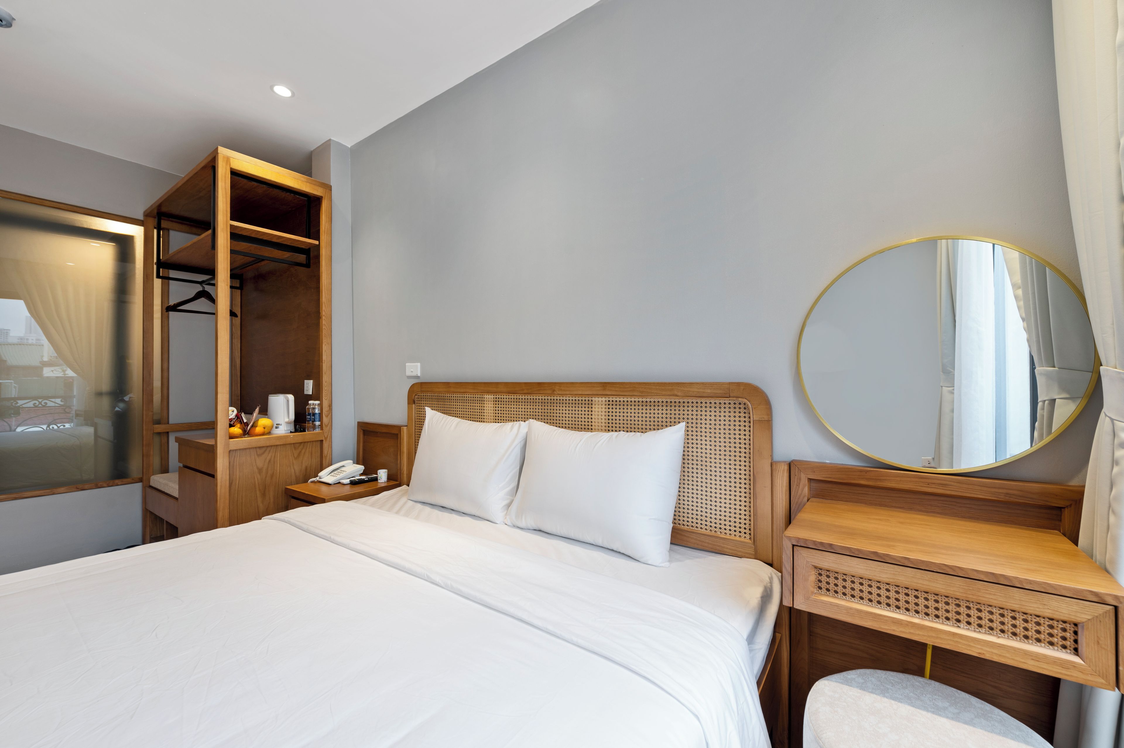 A hotel room with a bed, a wooden wardrobe, a bedside table with a phone, a round mirror, and a small seating area.