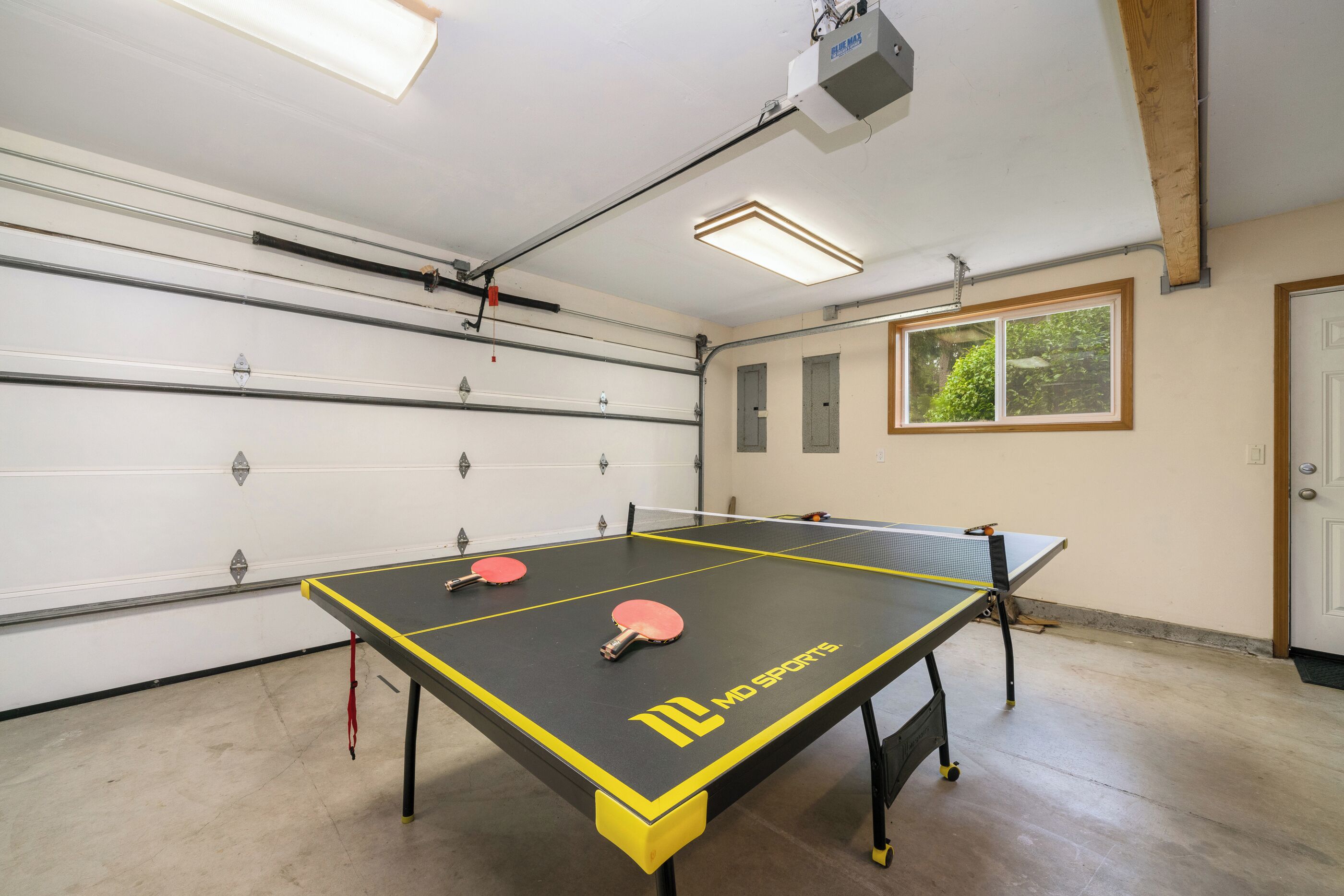 A ping pong table with two paddles, a ceiling-mounted projector, and a window with a view of greenery.