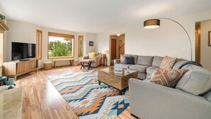 En rummelig stue med en stor sofa, et sofabord og et fjernsyn. Der er vinduer med rullegardiner, en lampe og et tæppe.