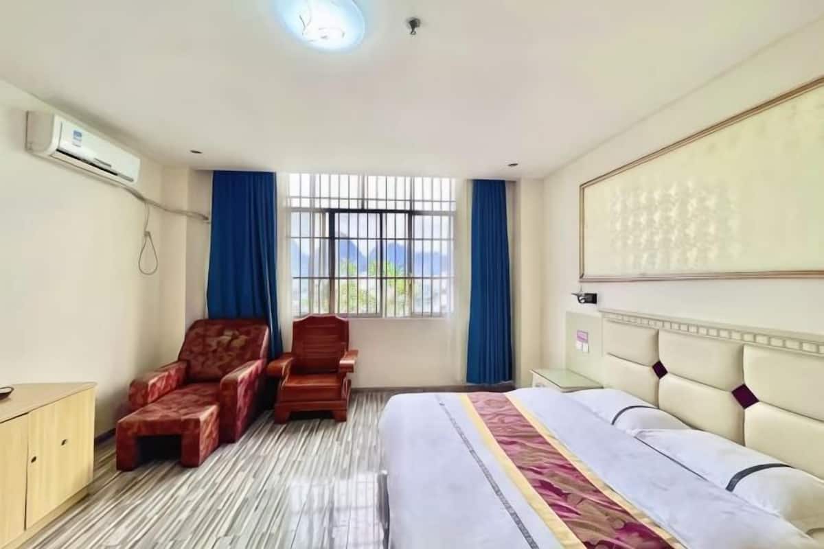 Hotellihuone, jossa on sänky, kaksi tuolia, työpöytä, ikkuna verhoilla ja seinään asennettu ilmastointilaite.