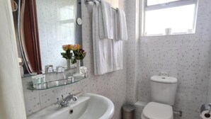 Ein Badezimmer mit weißem Waschbecken, Spiegel und Toilette. Handtücher hängen an einem Wäscheständer und ein Mülleimer steht neben der Toilette.