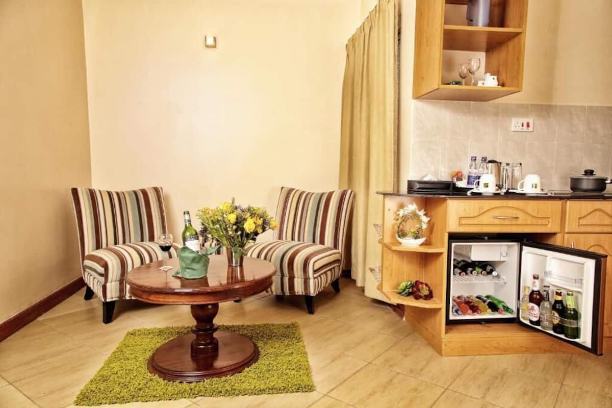 A compact living space with two striped armchairs, a round wooden table with a vase of flowers, a small kitchen area with a refrigerator, and a wooden cabinet above the counter.