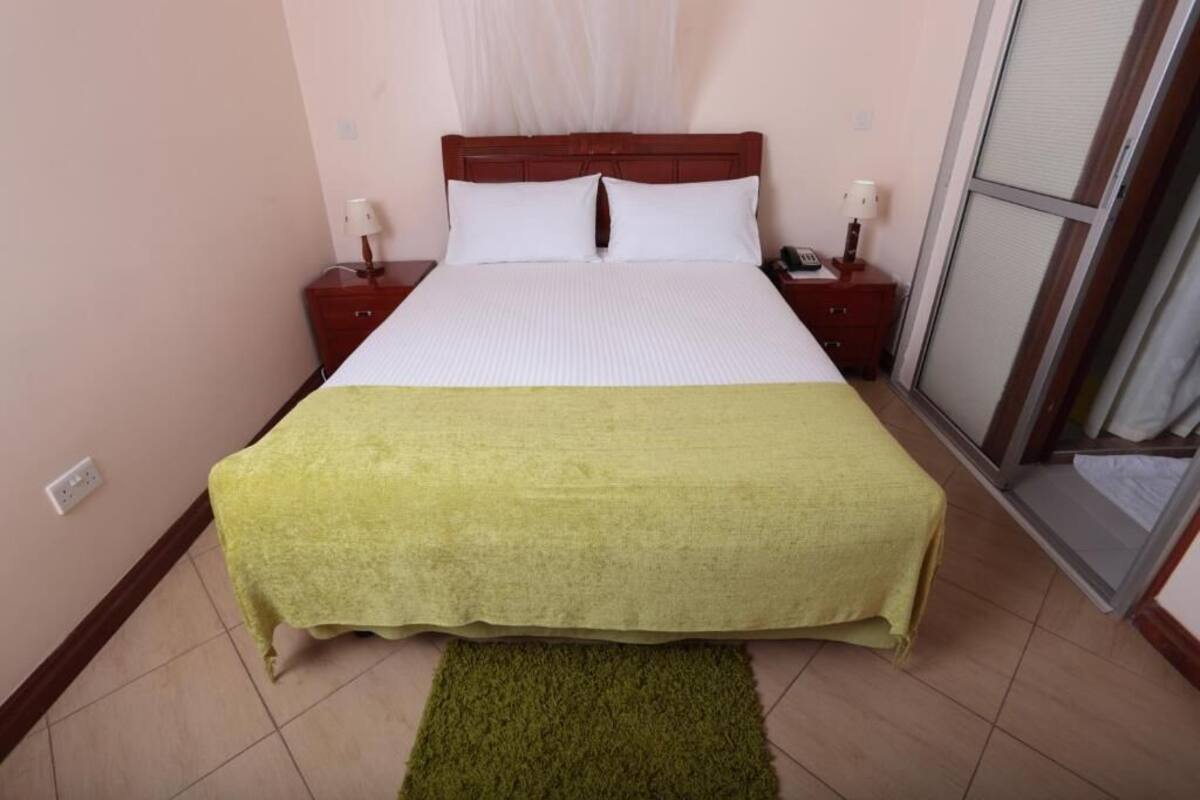 A neatly made bed with a white and green bedspread, two bedside tables with lamps, and a view of the outside through a sliding door.