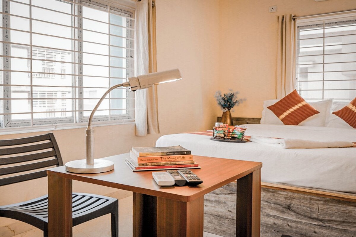 A hotel room with a bed, a bedside table with a lamp, a chair, and a window with blinds.