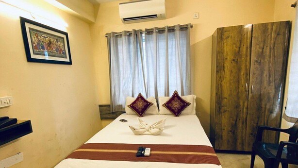 A hotel room with a bed, a ceiling fan, an air conditioner, a wooden wardrobe, a chair, and a framed picture on the wall. - Ostro Inn (Barasat)