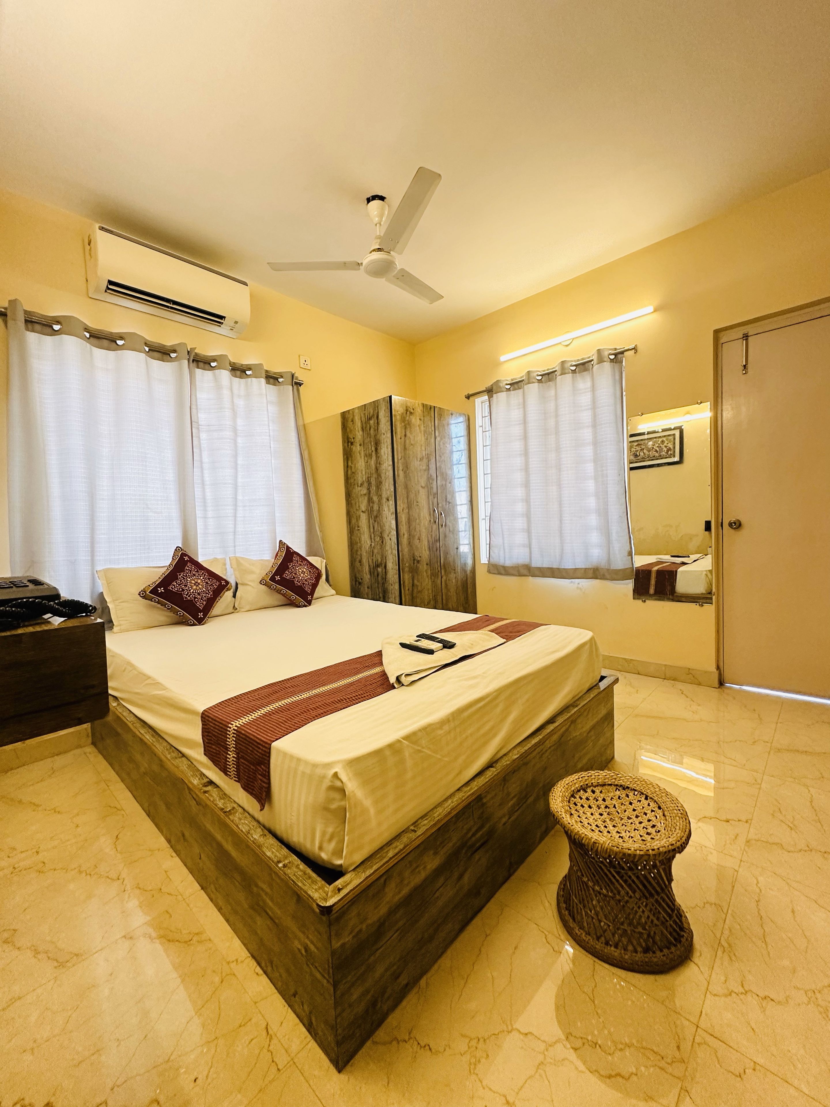 A hotel room with a wooden bed, a bedside table, a small stool, a ceiling fan, and a mirror on the wall.