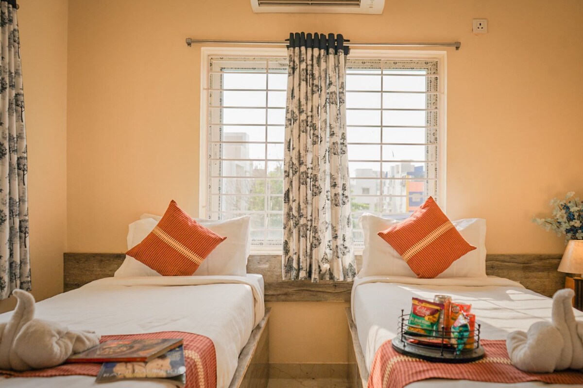 A hotel room with two beds, a window with floral curtains, a lamp, and a tray with snacks.