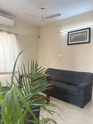 A living room with a grey sofa, a ceiling fan, an air conditioning unit, a framed picture on the wall, and a potted plant. - Ostro Inn (Barasat)